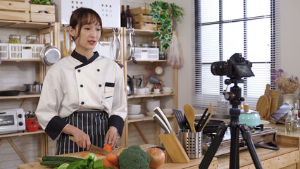 female chef vlogging at home kitchen. woman in chef’s uniform talking to camera while demonstrating different ways of cutting a carrot.