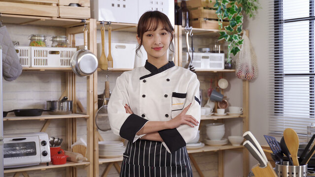 waist up, confident asian chef smiling at camera with arms crossed. background cozy home kitchen with wooden shelves. profession, profile picture concept