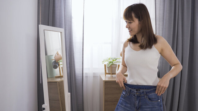 slim asian girl feeling happy about being able to fit into her jeans and standing in front mirror during clothes changing time in the morning.