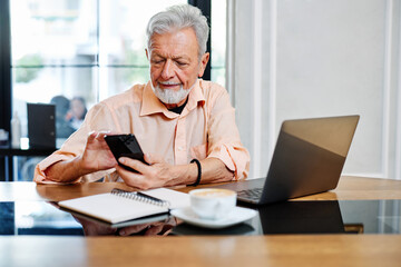 A senior student is sitting in a cafe and using his phone to check up on updates related to school. On the table are a laptop and notebook.