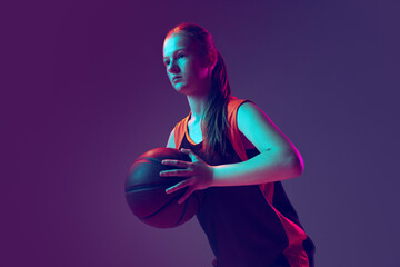 Naklejka premium Portrait of young girl, basketball player in brown uniform training isolated over gradient pink purple background in neon.