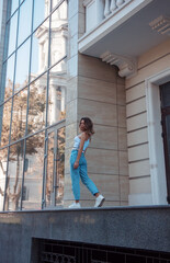 young curly brunette girl in white top and blue jeans is standing casual in step from the back and looking away near modern glass building background at city. lifestyle concept, free space