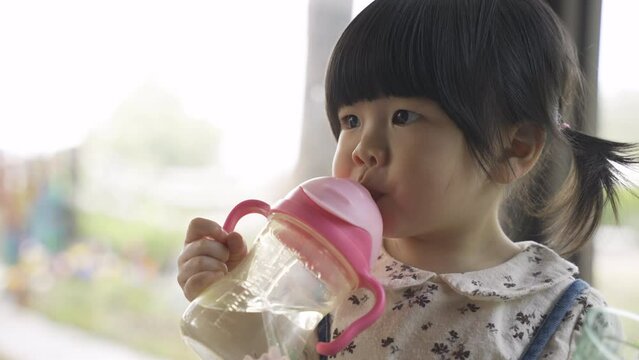 closeup shot of a lovely thirsty asian toddler girl holding a cup while drinking water from her sipper bottle in a bright restaurant.