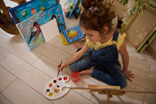 Overhead view of adorable little girl drawing and painting on canvas sitting on the floor next to wooden easel with beautiful drawn pictures at home. Kids art and children hobby, education concept