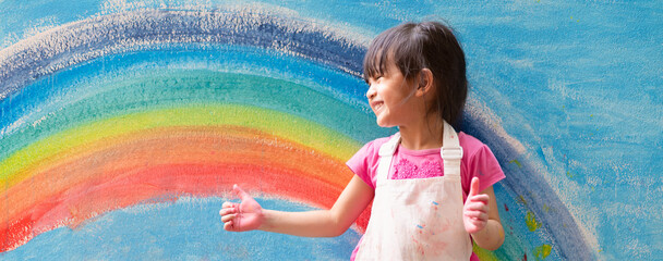sian little girl is painting the colorful rainbow and sky on the wall and she look happy and funny, concept of art education and learn through play activity for kid development.