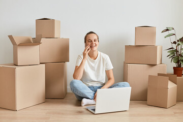 Optimistic beautiful woman wearing white t shirt sitting on floor surrounded with cardboard boxes with belongings and working on laptop, talking cell phone with positive emotions.
