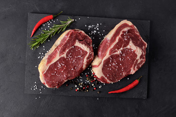 Two raw beef steaks on a cutting board with red hot peppers, rosemary and spices