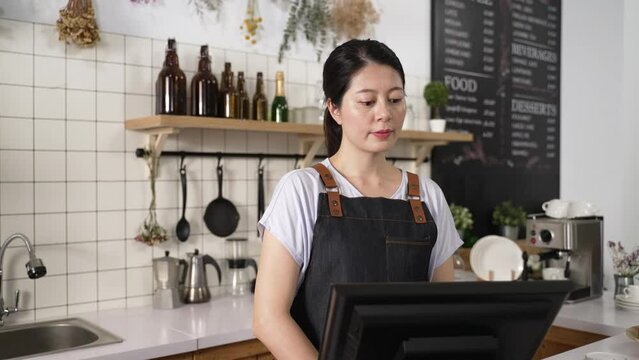 beautiful Asian coffee shop owner wearing apron looking up from monitor and smiling at the camera while operating a digital cashier machine.