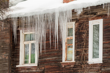 Multiple transparent icicles hang from the edge of the roof. Against the background of the wooden wall of the old house. Large cascades, even beautiful rows. Cloudy winter day, soft light. 