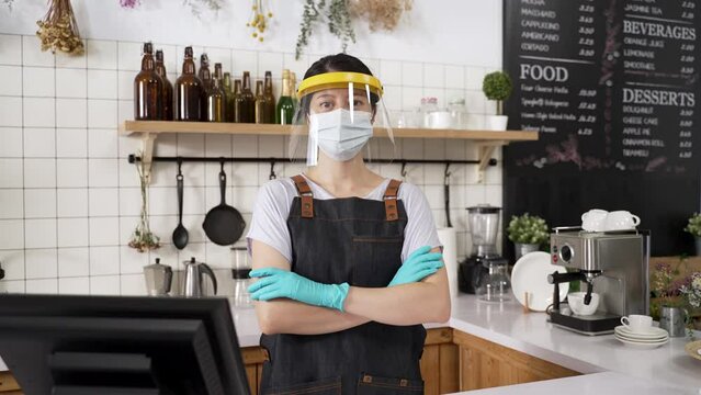 portrait of an asian coffee bar waitress wearing medical mask, gloves and plastic face shield is looking at camera with folded arms during pandemic outbreak.