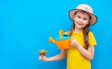Cute little gardener girl in a hat with gardening tools and in her hands on a blue background. The child is preparing to transplant a flower. The girl is a happy gardener