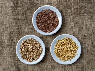 Flax seeds, raw wheat germ and buckwheat seeds on three ςηιτε plates on burlap fabric, top view