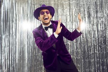 Happy goofy man wearing purple velvet suit, glasses and hat dancing, laughing and having fun at a party. Portrait of funny eccentric showman, toastmaster, or TV host on a silver foil fringe background - obrazy, fototapety, plakaty