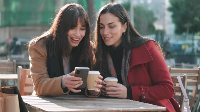 Video of wo attractive friends enjoying coffee together while looking smartphone and talking sitting on the terrace of a bar.