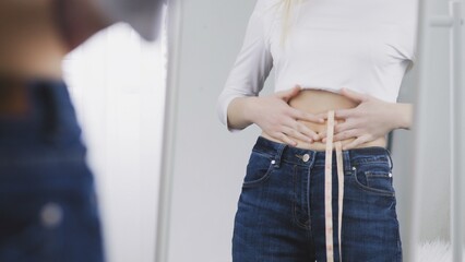 A young woman stands in front of a mirror and measures her waist circumference with a tape measure