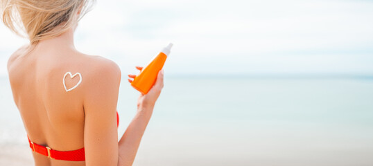Woman applying sun cream on tanned body In form heart love to protect her skin. Girl using sunscreen. Female holding suntan lotion, moisturizing sunblock on sunny day with blurry sea in the background - obrazy, fototapety, plakaty