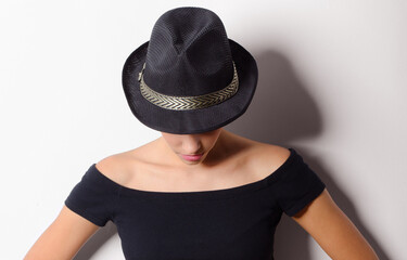 Beautiful fashionable woman in black clothes and black hat standing against white wall and looking down