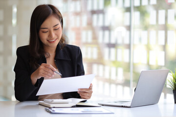 Smiling Asian businesswoman working on laptop and reading documents on desk in office.