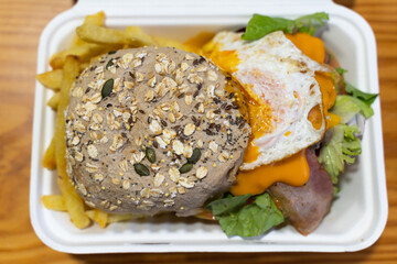 Big burger with seeds bread, fried egg, bacon, lettuce and french fries on take away container. Fast food to go concept