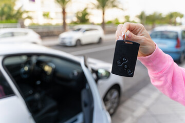 Hand of woman holding card key by white vehicle with open door. Car rental, free ride, insurance concepts