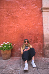 Caucasian woman smiling and checking her mobile phone while sitting on the floor held in a red wall on a medieval street.