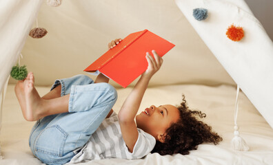 Cute little african american kid curly boy reading book in play tent - Powered by Adobe