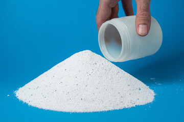 Laundry detergent on a blue background. A hand picks up washing powder in a measuring glass....