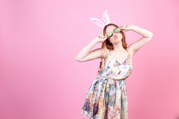 Obraz na płótnie Canvas Girl in a dress with rabbit ears on a pink background, Easter holidays. Woman beautiful with long hair, easter eggs closes her eyes.
