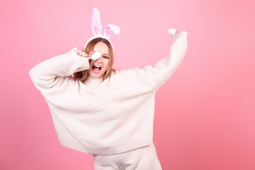 A girl with rabbit ears dances on a pink background. Easter holiday, smile and party. Woman posing in the studio. Closes eyes with easter eggs, having fun