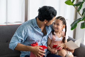 Selective focus at face of Father and Child Asian girl give a present to his dad while smile, hug and kiss inside of the living room. Happy moment with male parent, family relationship. Father day