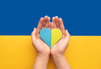 Stand with Ukraine concept. Hands holding blue and yellow heart on Ukraine flag background. Pray for Ukraine peace.
