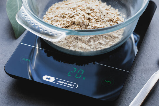 Oatmeal in a glass bowl on a kitchen scale on a dark gray background. Weighing the product. The concept of diet and calculation of proteins, fats and carbohydrates in a meal portion. Healthy eating