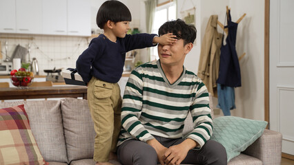 Fototapeta premium lovely Chinese baby boy hiding gift at back and covering dad’s eyes. surprised father embracing his son on receiving the father’s day present in the living room at home