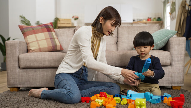 happy asian mother and baby son sitting on ground playing toy blocks together in the living room at home. she helps give him parts to build