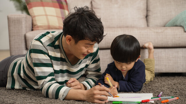 loving asian father talking to his cute baby son while lying on belly on floor drawing pictures with him. love and family concept
