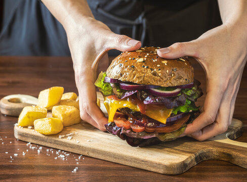Close-up of a delicious burger with vegetables and fries on a dark wooden table.An unrecognizable person picks up the burger with two hands to eat it.
