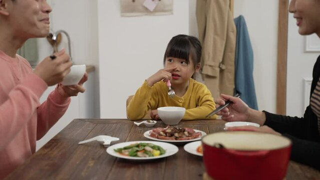 selective focus of picky Asian little girl looking at her father eating vegetable with hesitation. her mother helps her pick up meat dropping from the fork at dining table