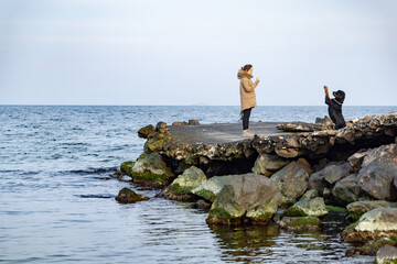 Fototapeta na wymiar A girl in a jacket walks on the coast of the sea with a Rottweiler dog