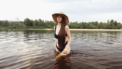 A young brunette girl in a hat stands in the water. Girl with long hair in a black bathing suit. Swimming in nature in summer