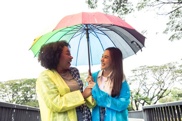 Rainy day, Positive young diversity woman wearing yellow and blue raincoat during the rain in the park. Dodge and run away from the rain with colorful umbrella in a joyful manner.