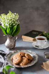 English 5 o'clock tea. Oat cookies or biscuits on a vintage plate, a cup of black tea, books and may-lily bouquet on dark background. Greeting card design, cafe, bakery poster