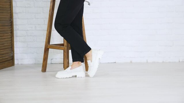 Fashionable white shoes presentation. Female model walks in fashionable footwear back and forth near the wooden tabouret.