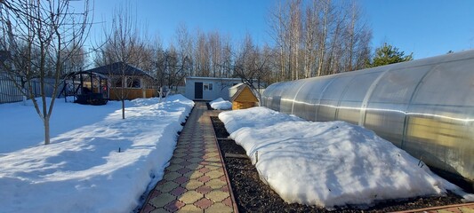 Sunny clear day in early spring. View of a suburban area with greenhouses, a well and a barn. Garden paths without snow, snowdrifts on flower beds and beds