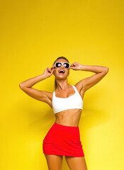 young blonde fit girl in white top and red tennis skirt is standing casual on the yellow wall background and laughing happy with hands near sunglasses. sport lifestyle concept, free space