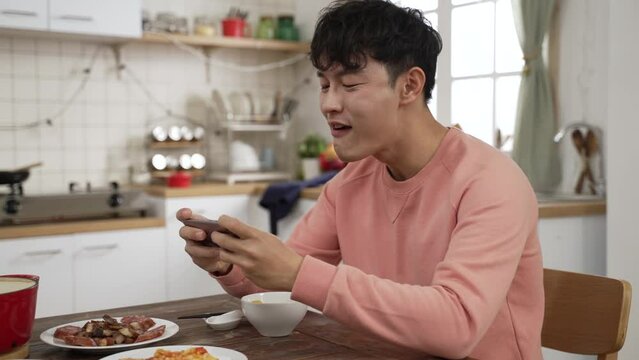 distracted asian father playing mobile game with smartphone while having meal at dining table. he cheers for victory with clenched fist and takes a bite of meat
