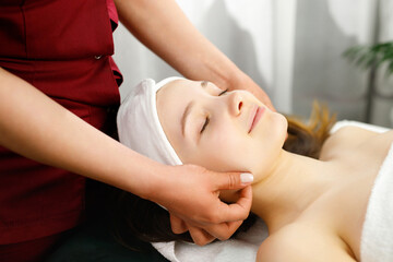 Fototapeta na wymiar Calm and relaxed woman with closed eyes undergoing face massage being in spa salon by a masseur in red uniform.