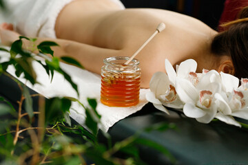 Young woman lying on the massage table with face down covered with towel and jar of honey near the...