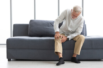 senior man suffering from knee ache on sofa - obrazy, fototapety, plakaty