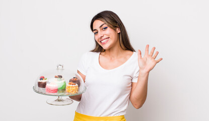 pretty hispanic woman smiling happily, waving hand, welcoming and greeting you. home made cakes concept