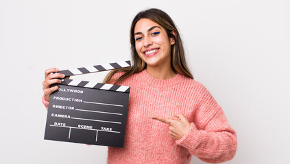 pretty hispanic woman smiling cheerfully, feeling happy and pointing to the side. cinema clapper...